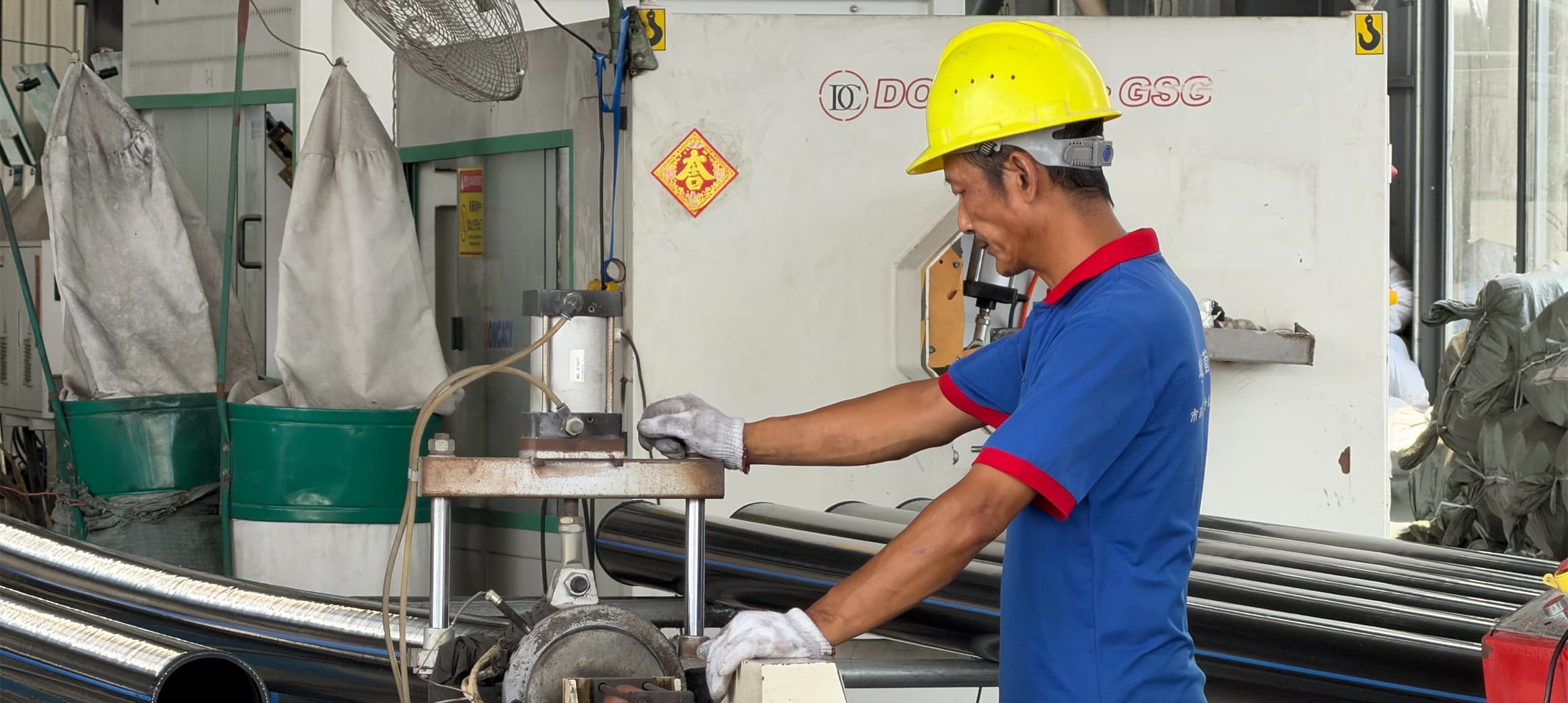 A worker in a hard hat and safety gloves guiding black PE100 pipe through the cut-off machine on the workshop floor